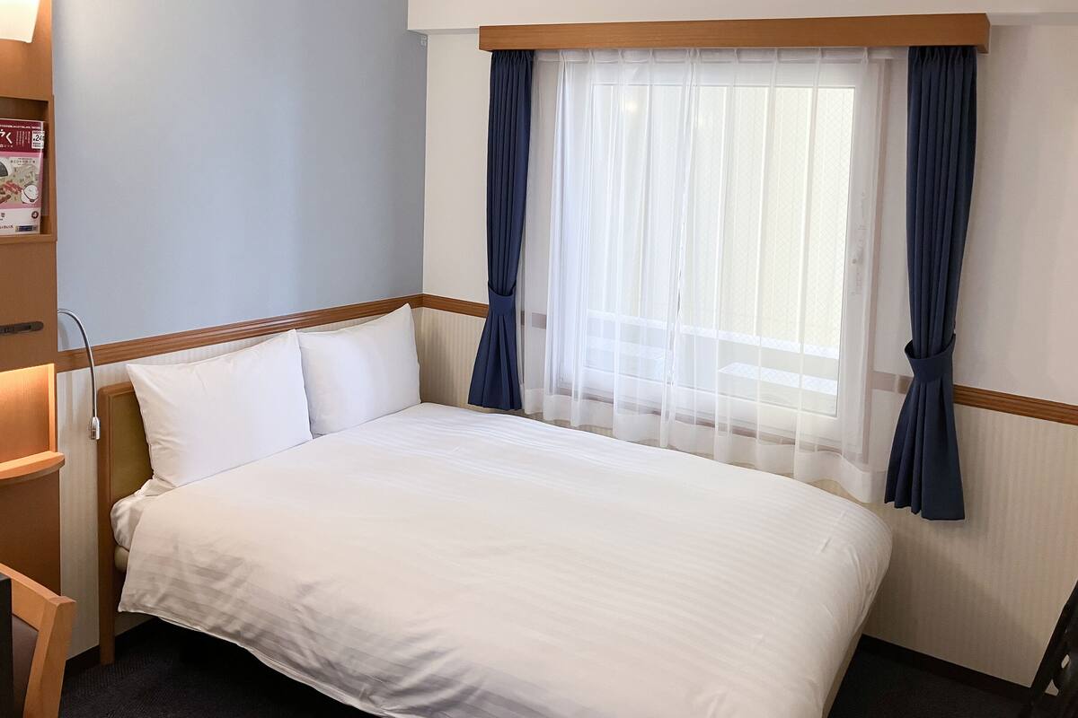 Une chambre d’hôtel comprenant un lit, une table de chevet, une lampe, une fenêtre avec des rideaux et une horloge murale.