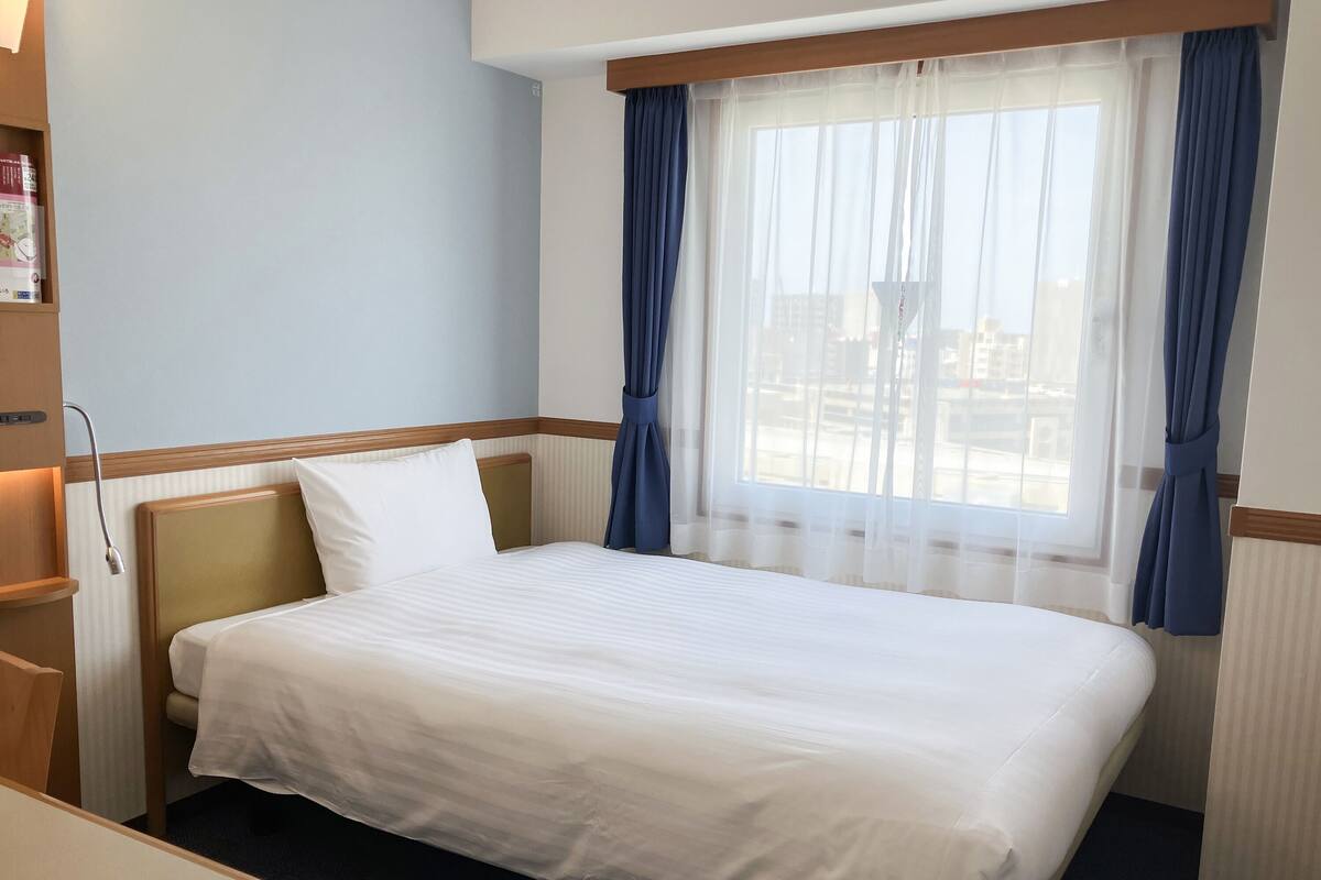 Une chambre d’hôtel avec un lit, un bureau, une bibliothèque et une fenêtre avec des rideaux.