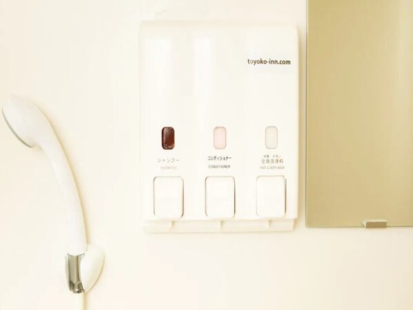 Een witte hotelkamer met een bedtelefoon, een klein nachtkastje en een bedieningspaneel aan de muur met drie knoppen, gelabeld 'Shampoo', 'Conditioner' en 'Gezicht & Lichaamswas'.