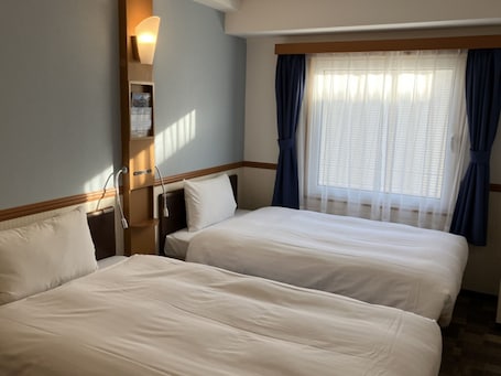 A hotel room with two beds, a bedside table, a lamp, and a window with curtains.
