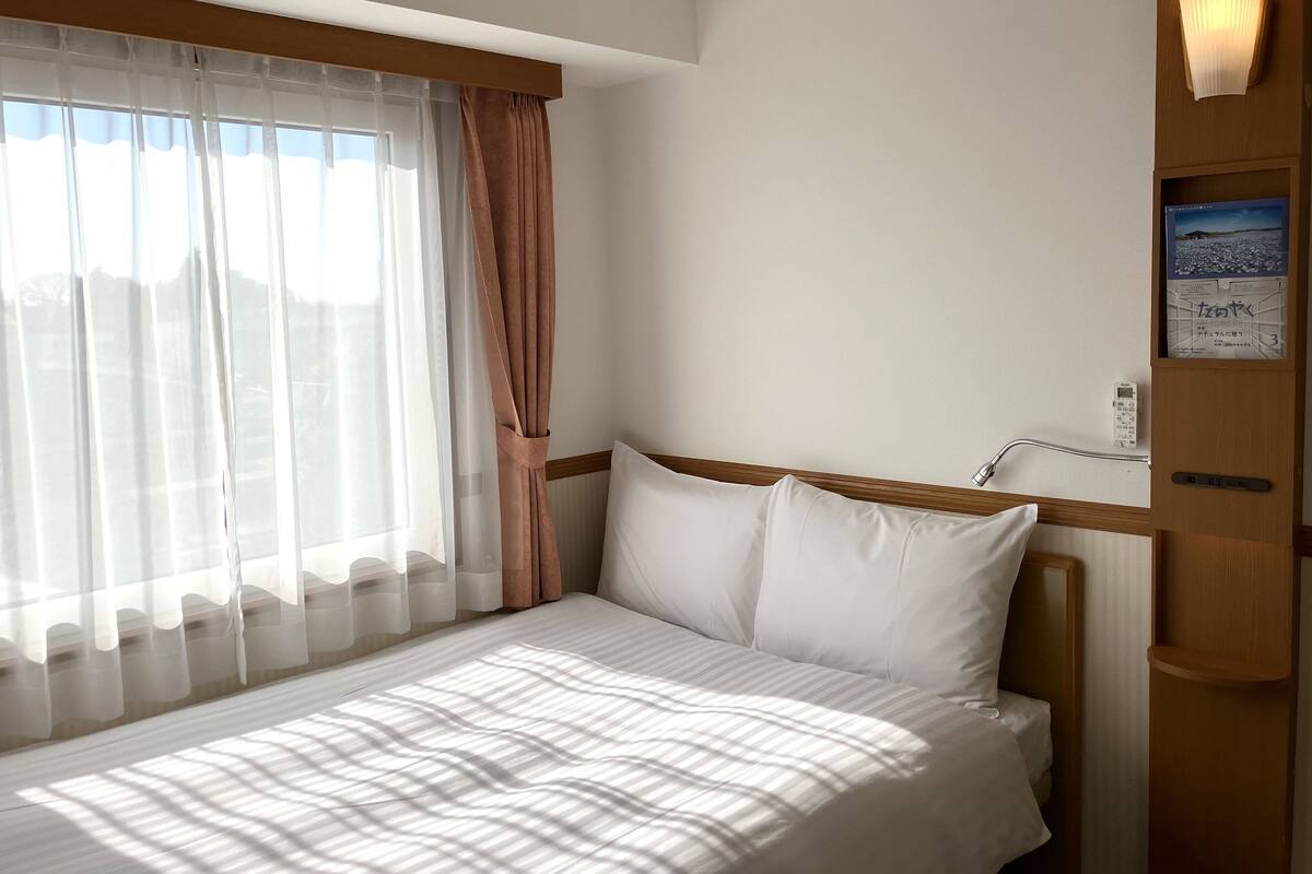 A hotel room with a bed, a bedside table, a lamp, and a window with curtains.