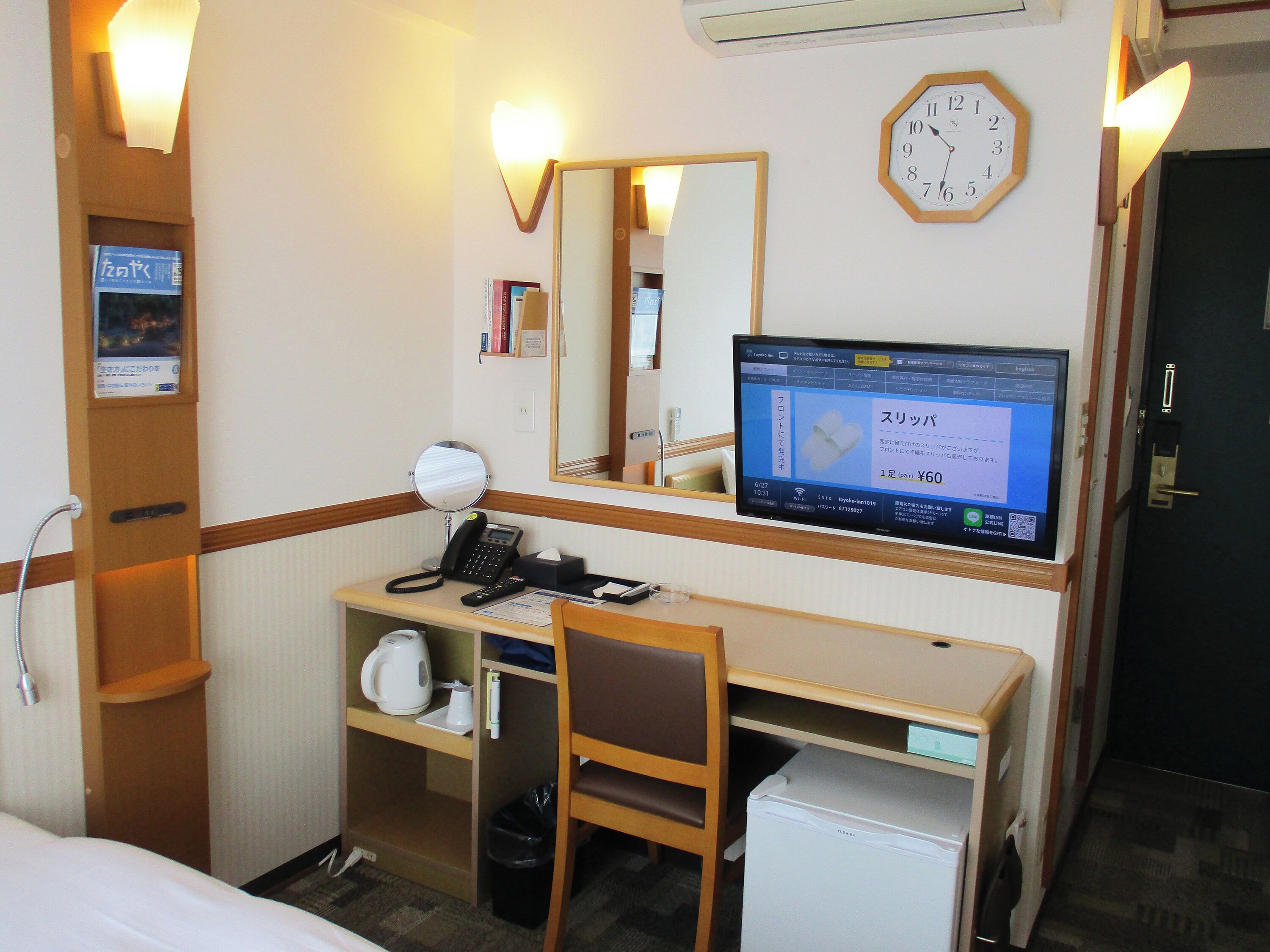 A hotel room with a desk, chair, bed, bedside table, mirror, clock, and a television displaying a message in Japanese.