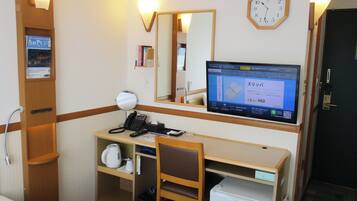 A hotel room with a desk, chair, bed, bedside table, mirror, clock, and a television displaying a message in Japanese.