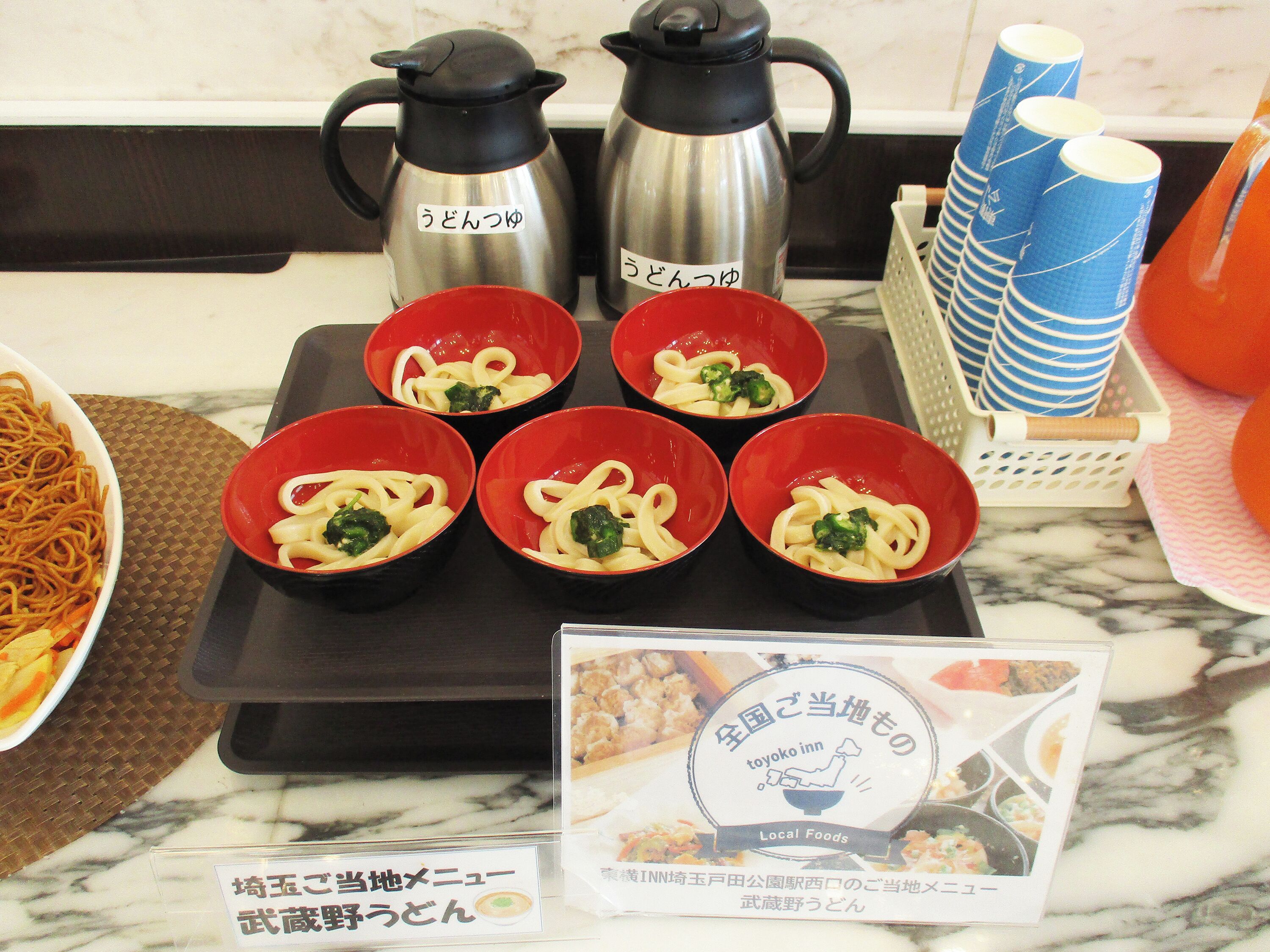 A kitchen counter with four red bowls of pasta, two stainless steel coffee pots, and a basket of blue and white striped cups.