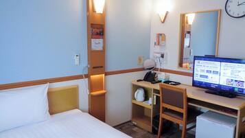 A hotel room with a bed, a desk with a computer, a chair, a small refrigerator, and a wall-mounted mirror.