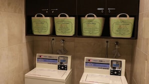 Two front-loading washing machines with digital displays, a trash bin, and green laundry bags on a shelf above. - Toyoko Inn Tokushima Ekimae (Tokushima)