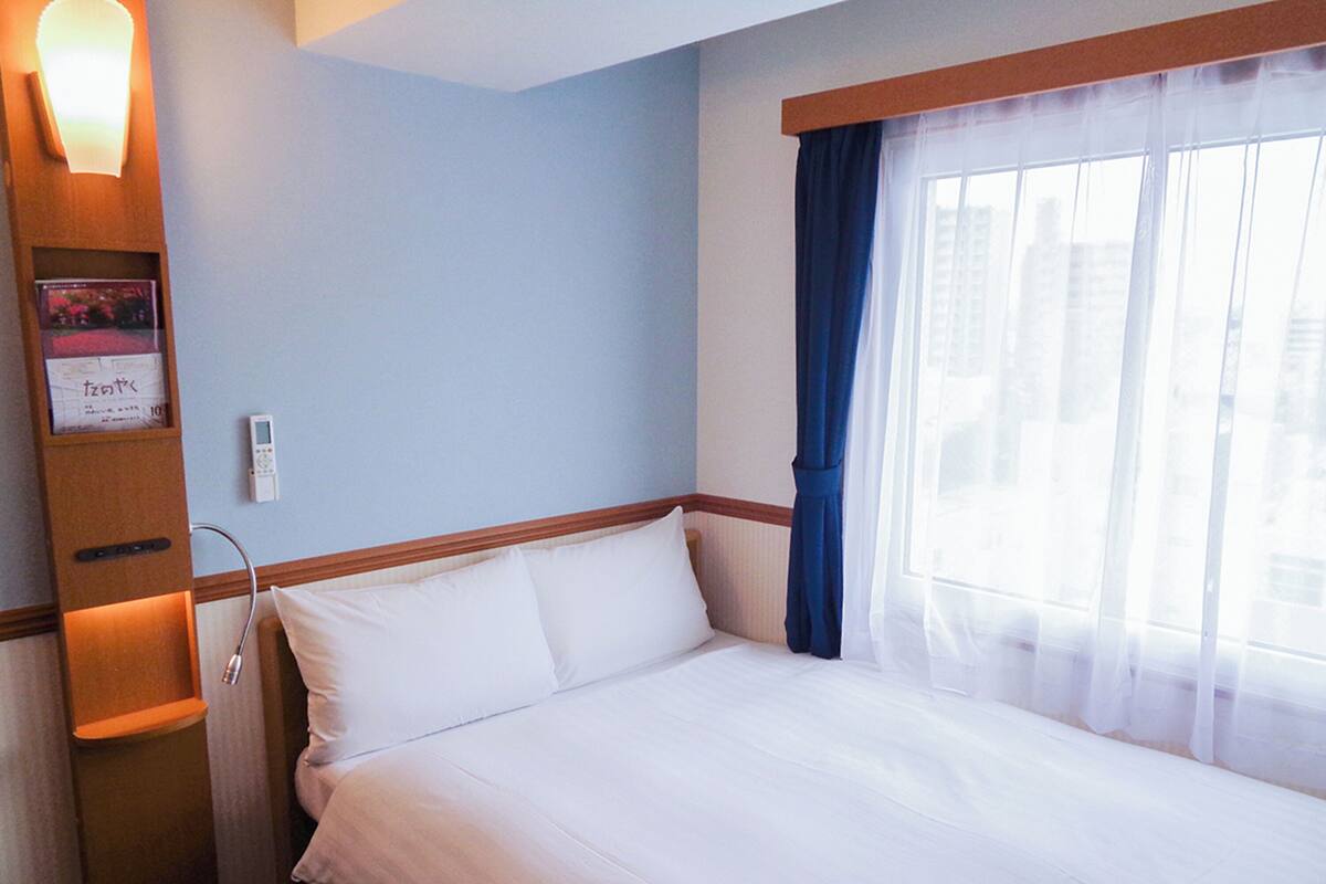A hotel room with a bed, a wall-mounted control panel, and a window with curtains.