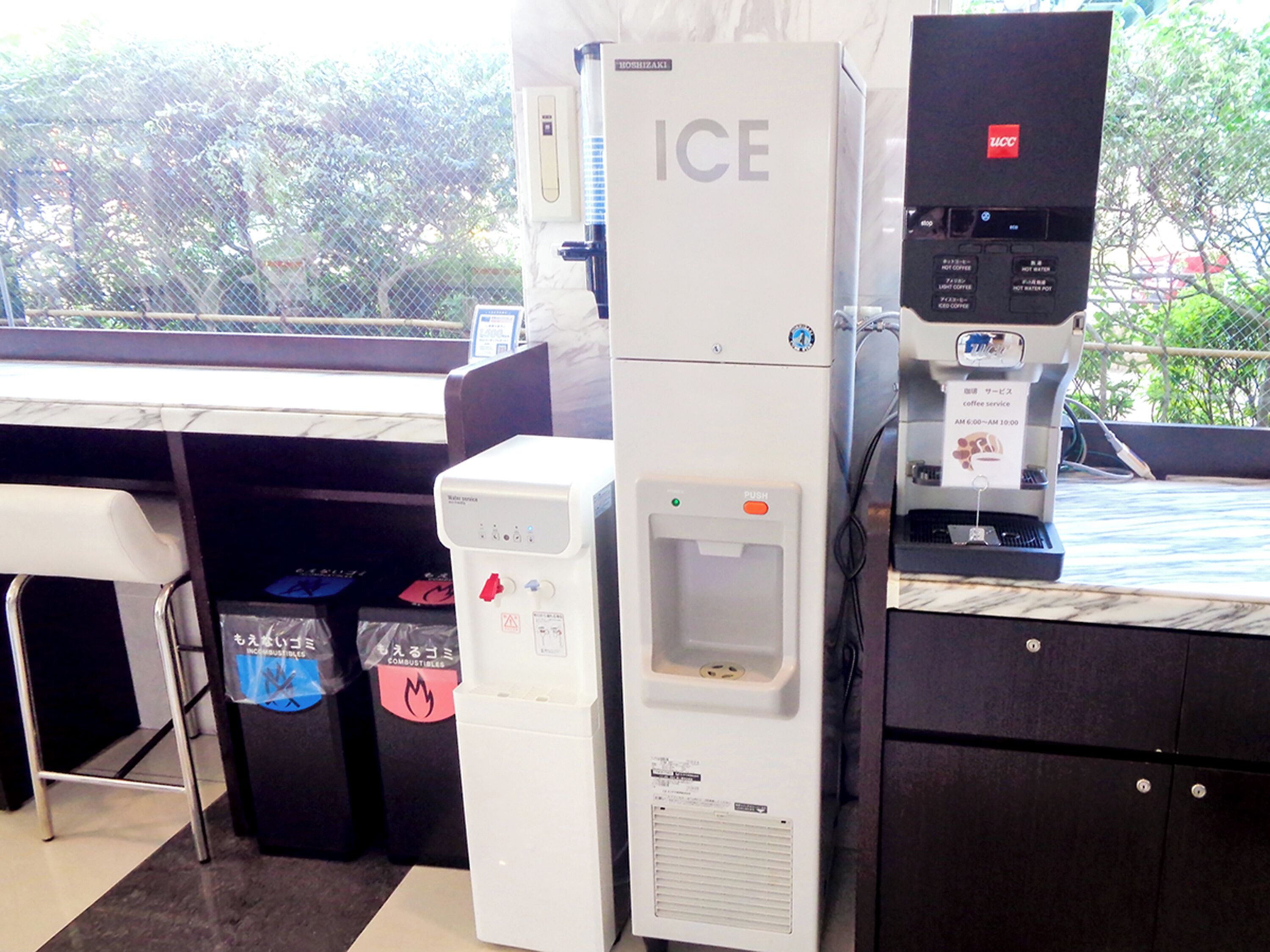 A coffee machine and a water dispenser in a public area, with trash bins and a seating area visible.