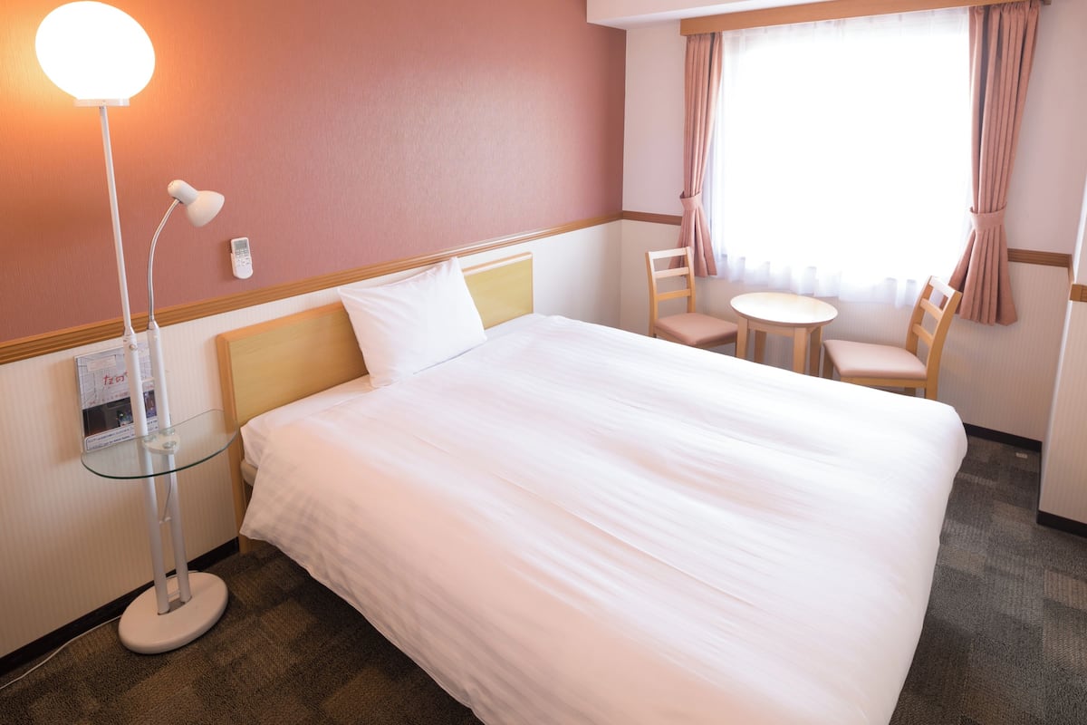 Ett hotellrum med en sÀng, en sÀnglampa, en stol, ett bord och ett fönster med gardiner.
