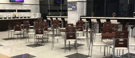 A spacious dining area with multiple tables and chairs, a television mounted on the wall, and a clean, tiled floor.