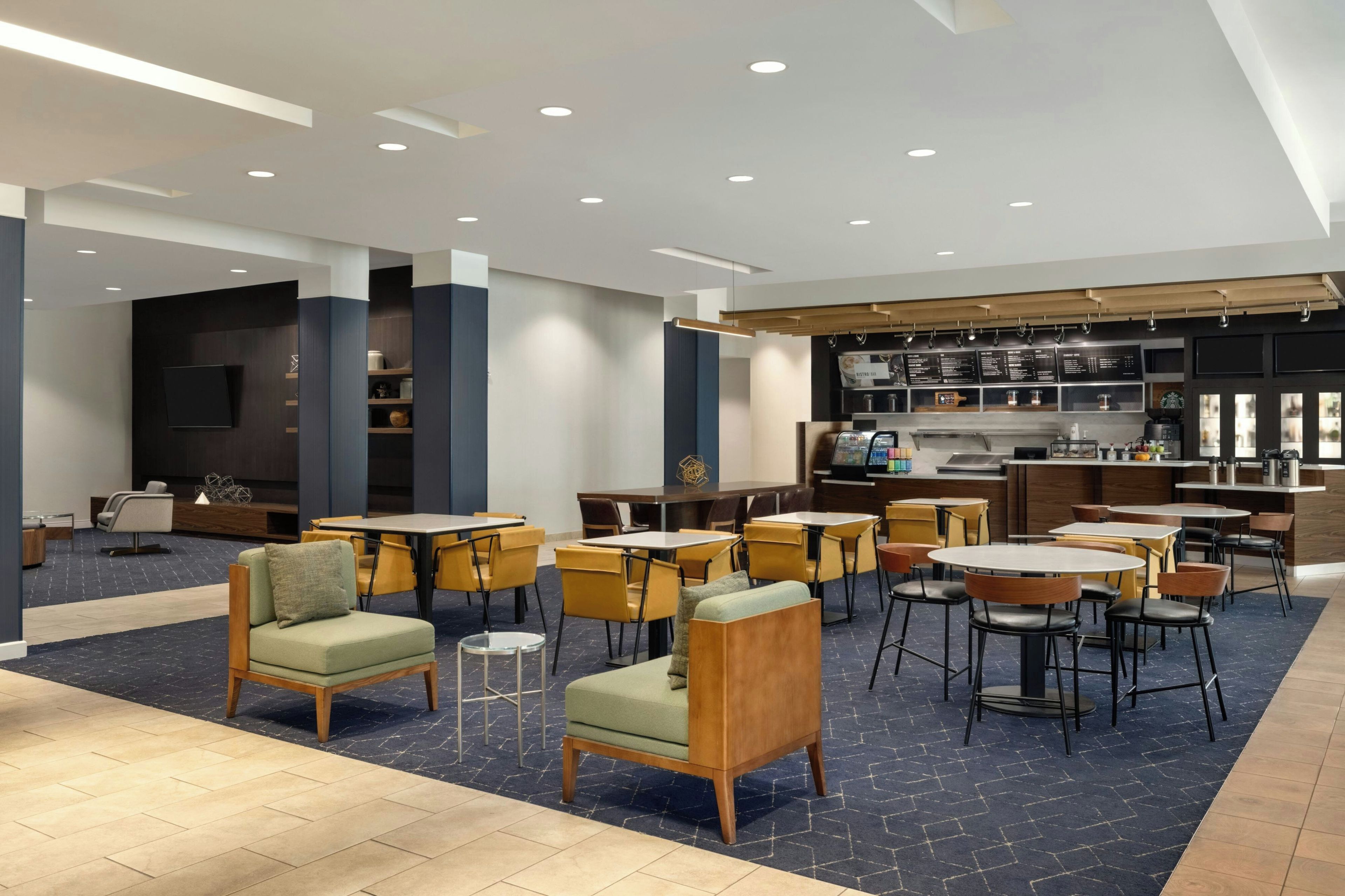 Un hall d’hôtel moderne doté de plusieurs espaces de détente, d’un bar et d’une cuisine visible au fond.