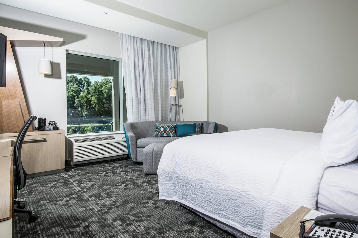Habitación de hotel con cama, escritorio, silla, lámpara y vista a los árboles por la ventana.