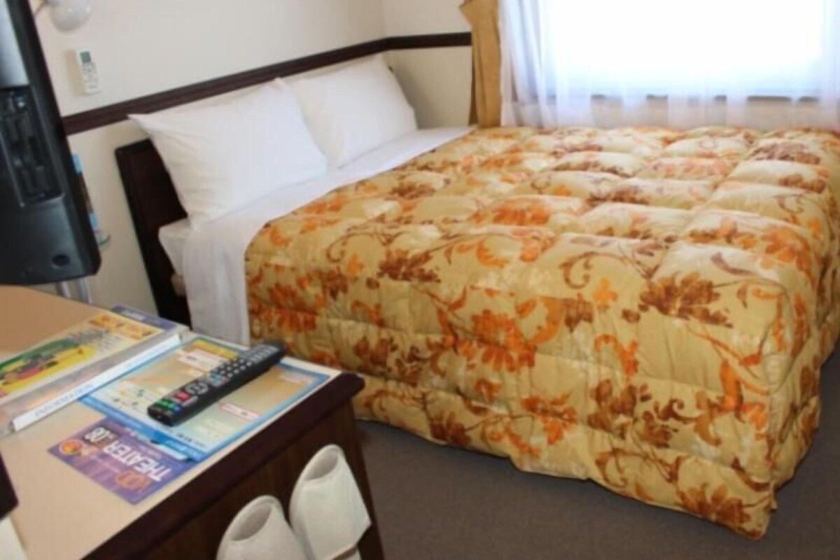 A hotel room with a bed, a desk with a remote control, and a pair of slippers.