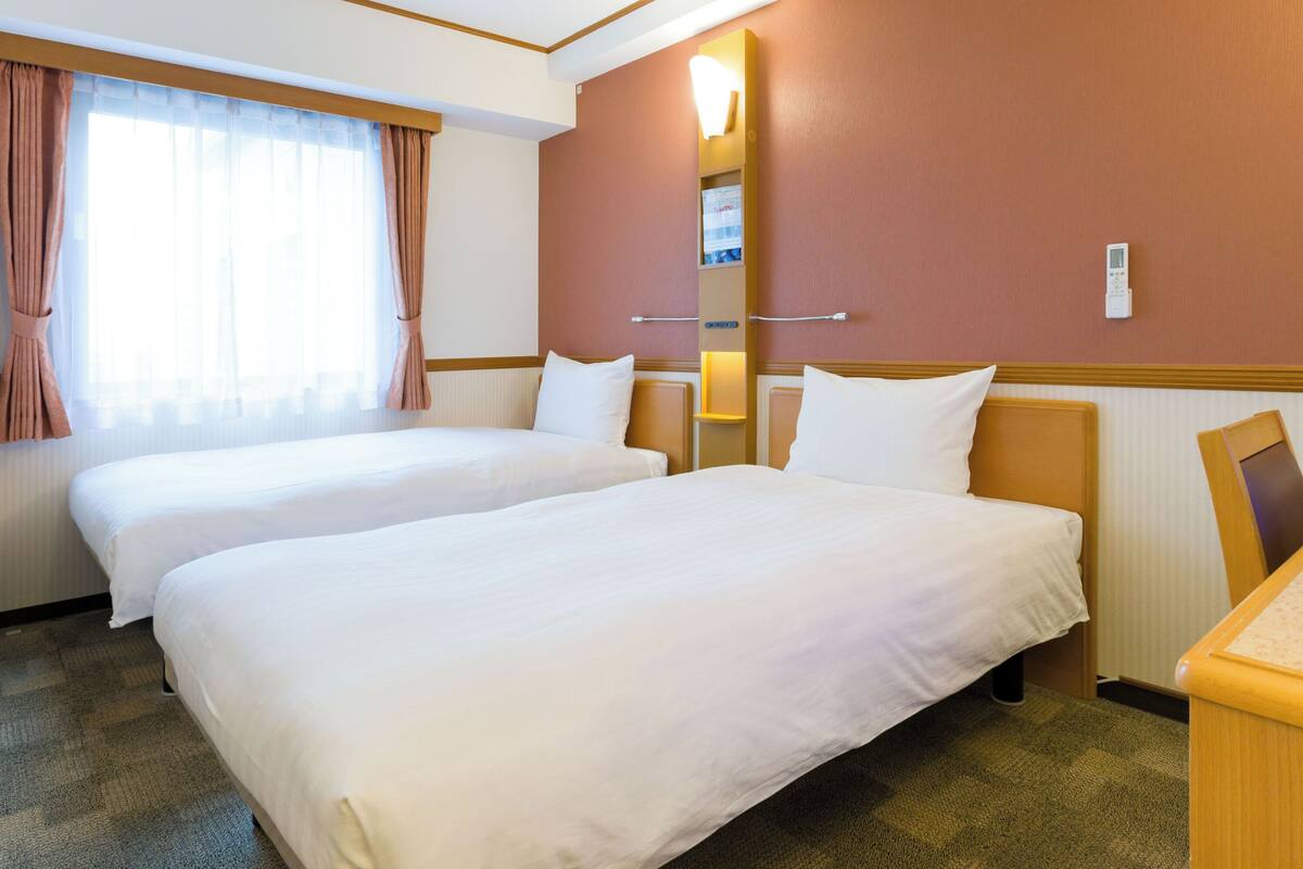 Ein Hotelzimmer mit zwei Betten, einem großen Fenster mit Vorhängen, einer Wandlampe und einem kleinen Schreibtisch.