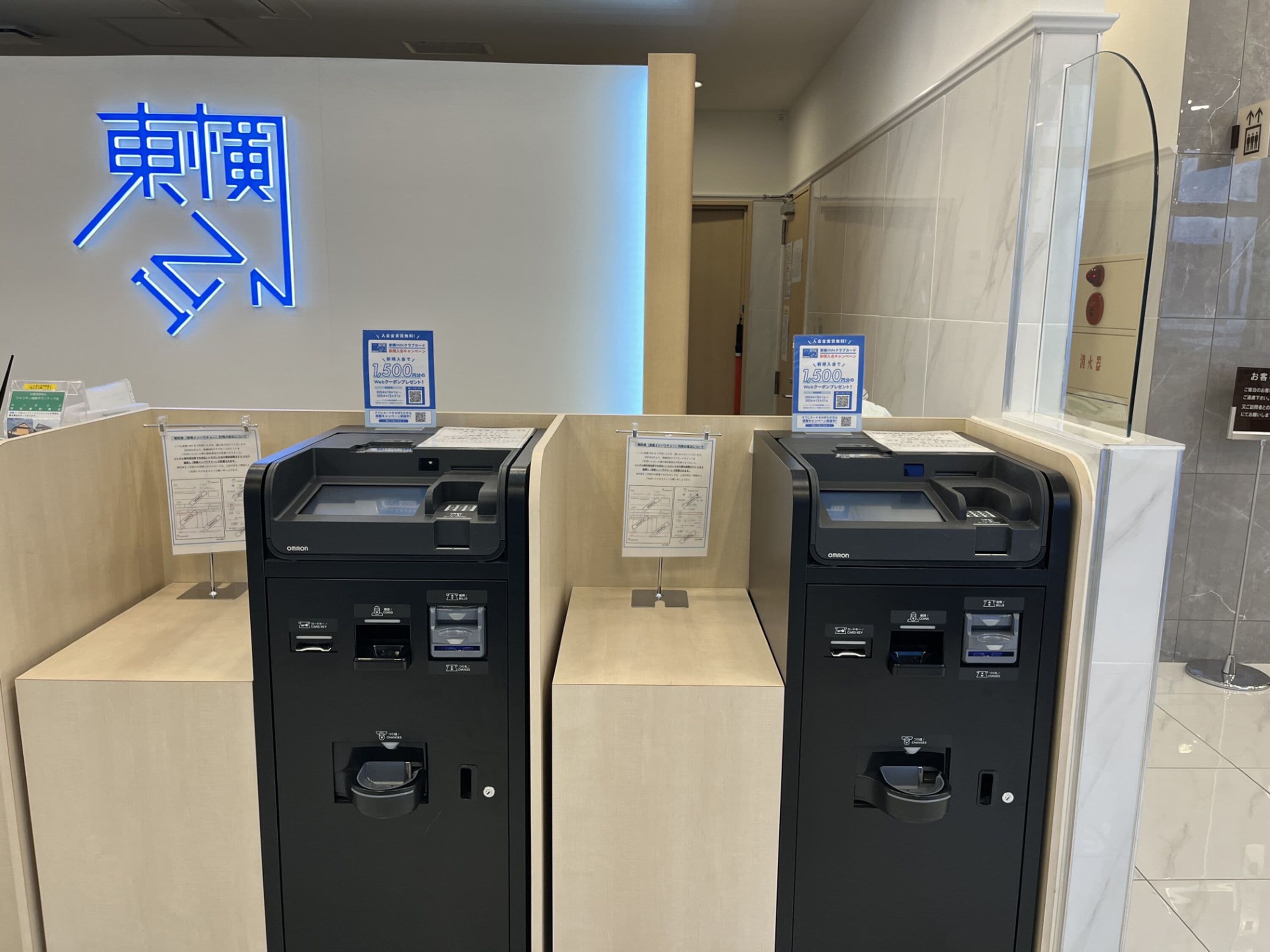 A modern hotel reception area with two Omron self-service kiosks, a sign with directional arrows, and a mirror on the wall.