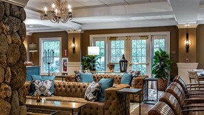 Un salon spacieux avec un mur d’accent en pierre, un grand canapé en cuir marron, une table basse en bois et un fauteuil en osier. Il y a deux fenêtres avec des cadres blancs et un lustre suspendu au plafond.