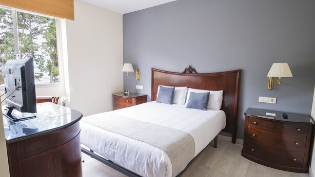 Une chambre d’hôtel comprenant un lit, des tables de chevet, un bureau avec un ordinateur et une fenêtre donnant sur des arbres.