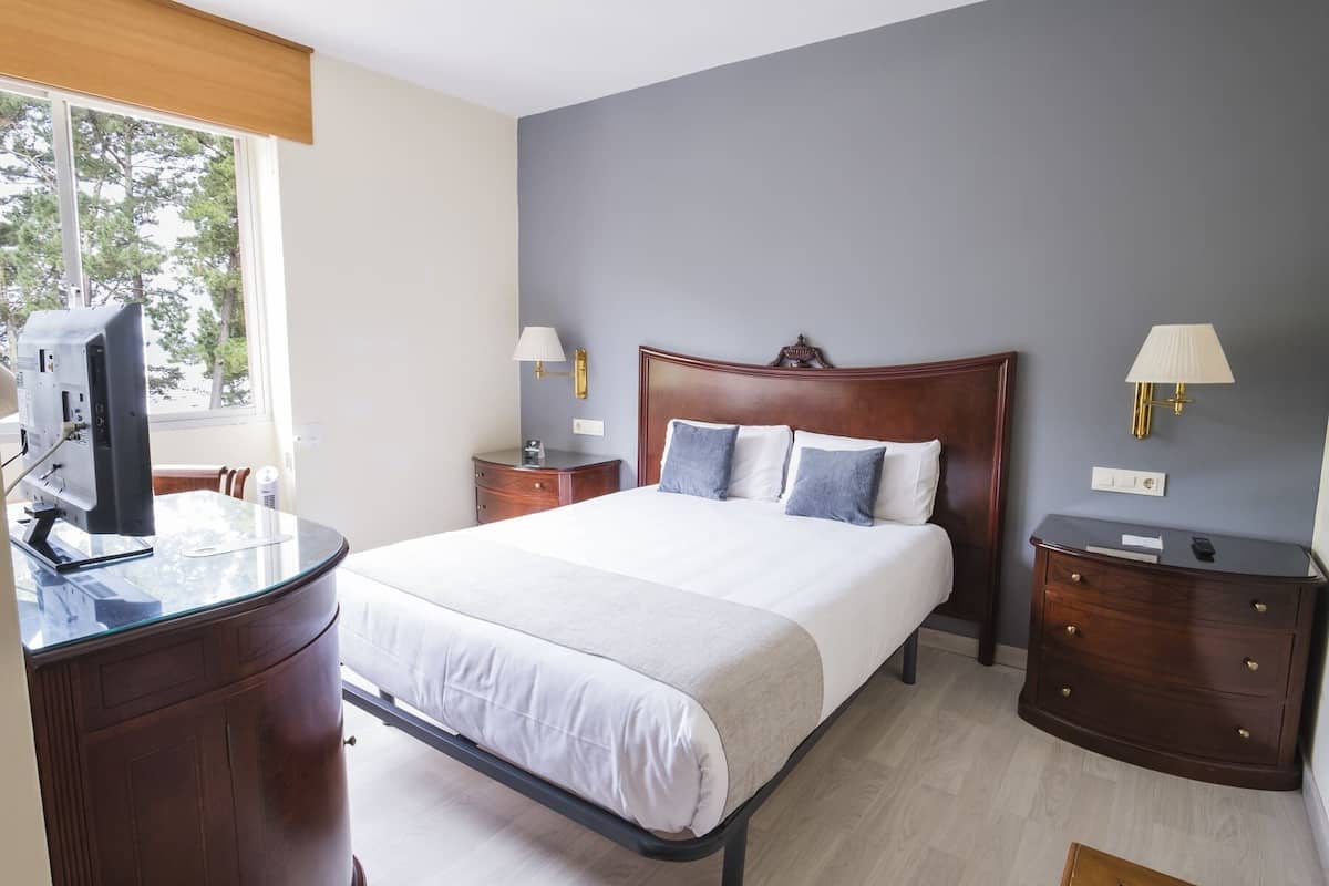 A hotel room with a bed, bedside tables, a desk with a computer, and a window with a view of trees.