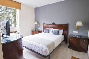 A hotel room with a bed, bedside tables, a desk with a computer, and a window with a view of trees. - Alda Cabanas Playa (Cabanas)