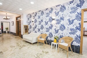 Un vestíbulo con un sofá blanco, dos sillones de mimbre, una mesita con un florero y un papel tapiz estampado con motivos florales.