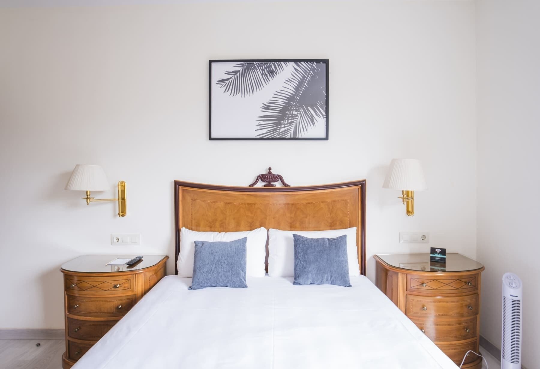 Une chambre d’hôtel avec une tête de lit en bois, deux tables de chevet avec des lampes, une œuvre d’art représentant une feuille encadrée, et un lit avec une literie blanche et deux oreillers.