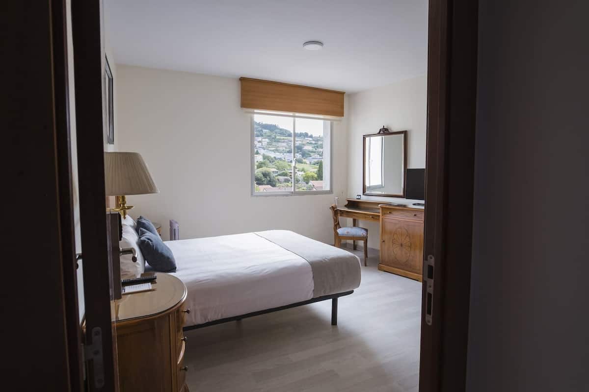 A bedroom with a bed, a desk, a chair, a lamp, a mirror, and a window with a view of a town.