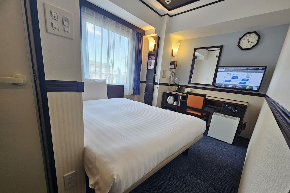 Hotelkamer met een bed, een bureau, een stoel, een kleine koelkast, een klok en een digitaal scherm.