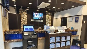 Zona de recepción de hotel con pantalla digital, quiosco de autoservicio y mostrador con toallas dobladas.