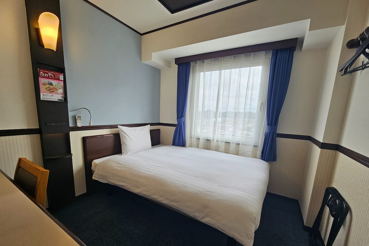 Hotelkamer met een bed, bureau, stoel, nachtkastje, lamp en raam met gordijnen.