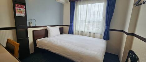 A hotel room with a bed, a desk, a chair, a nightstand, a lamp, and a window with curtains.