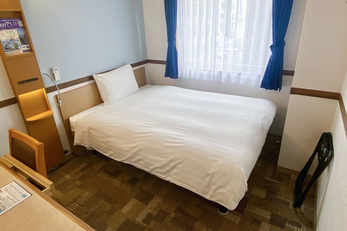 Una habitación de hotel con cama, escritorio, silla, estante con libros y ventana con cortinas.