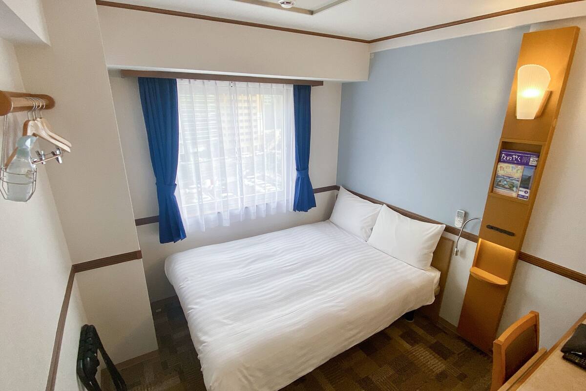 Habitación de hotel con cama, ventana con cortinas azules, escritorio con silla y lámpara fijada a la pared.