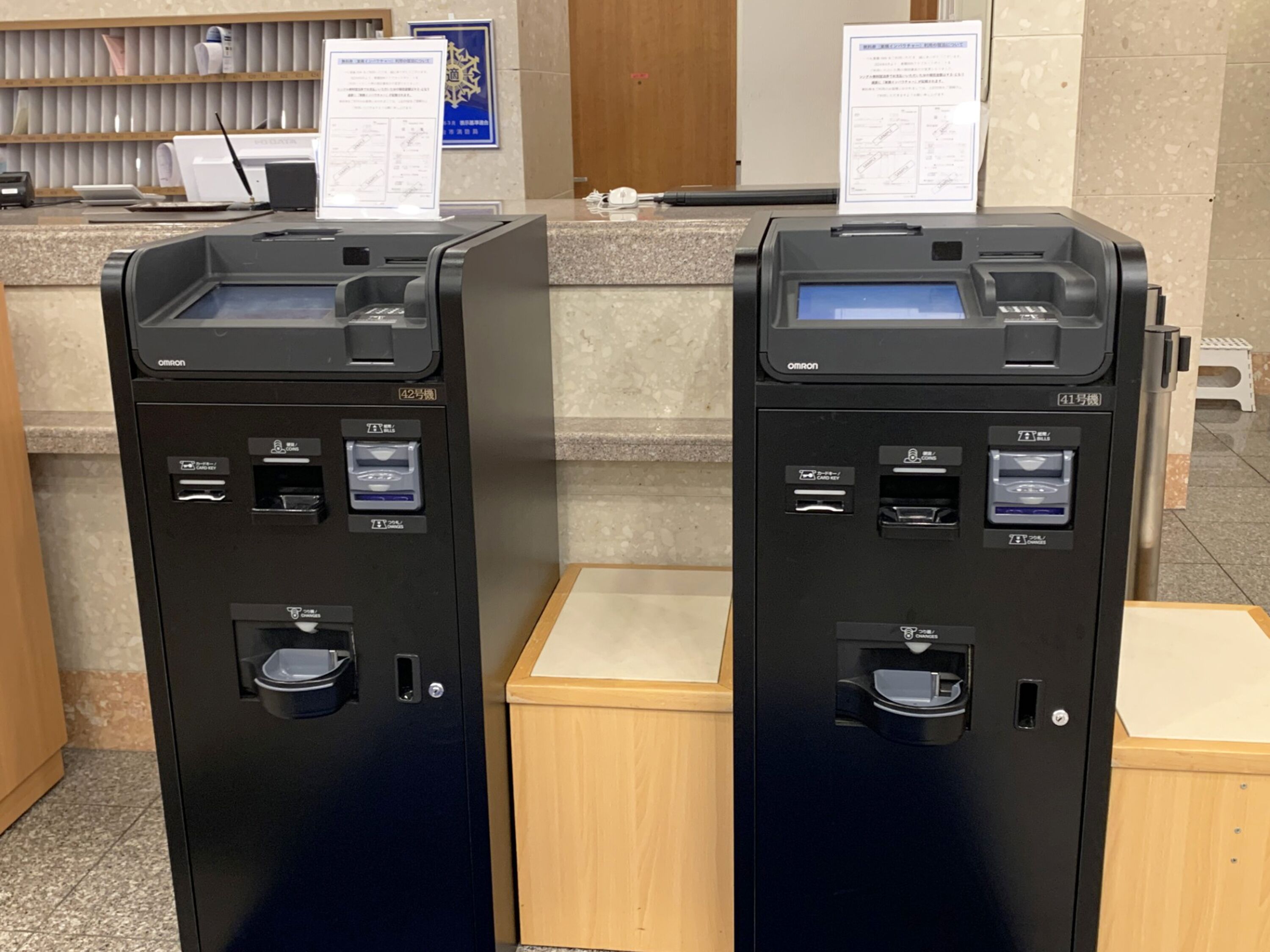 Two Omron self-service kiosks with card readers, a small trash bin, and a wooden counter.