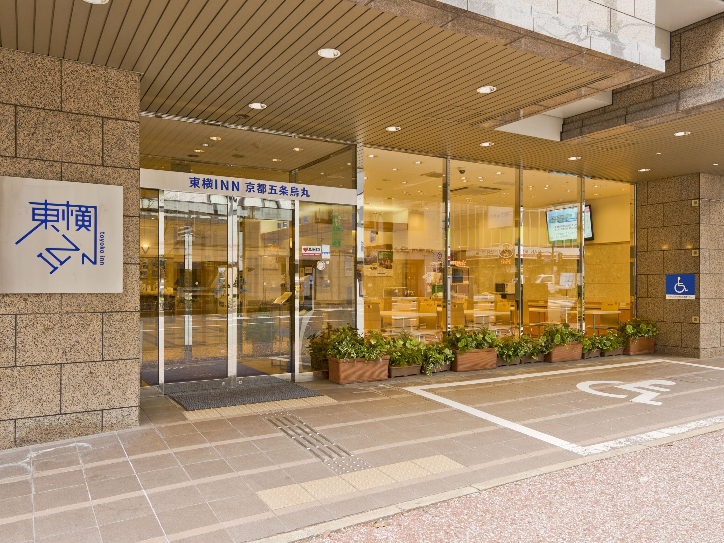 L’entrée d’un hôtel, avec une enseigne indiquant « Toyoko Inn » et le symbole d’un défibrillateur automatisé externe (DAE).