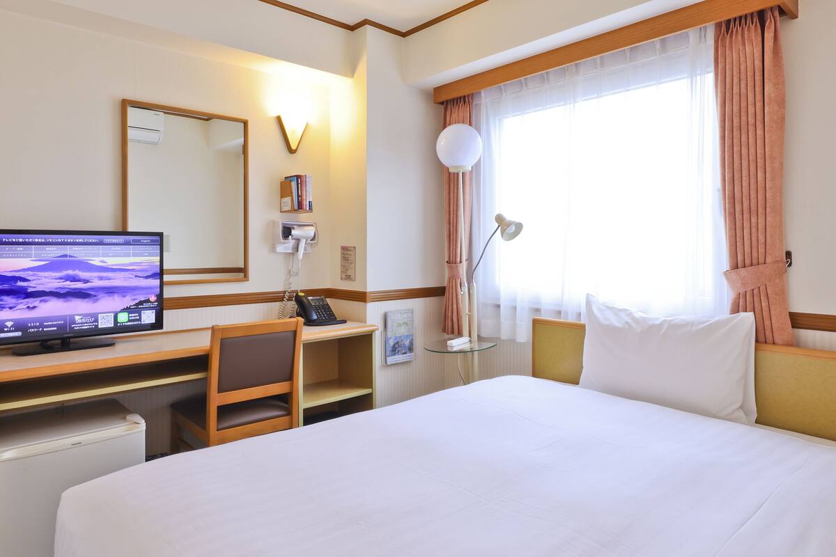 A hotel room with a bed, desk, chair, TV, phone, lamp, and window with curtains.
