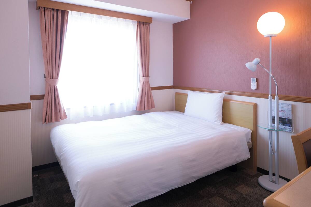 A hotel room with a bed, a bedside lamp, a window with curtains, and a wall-mounted telephone.