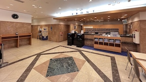 A hotel reception area with a clock, a reception desk, a counter with a computer, and a seating area with a bench.