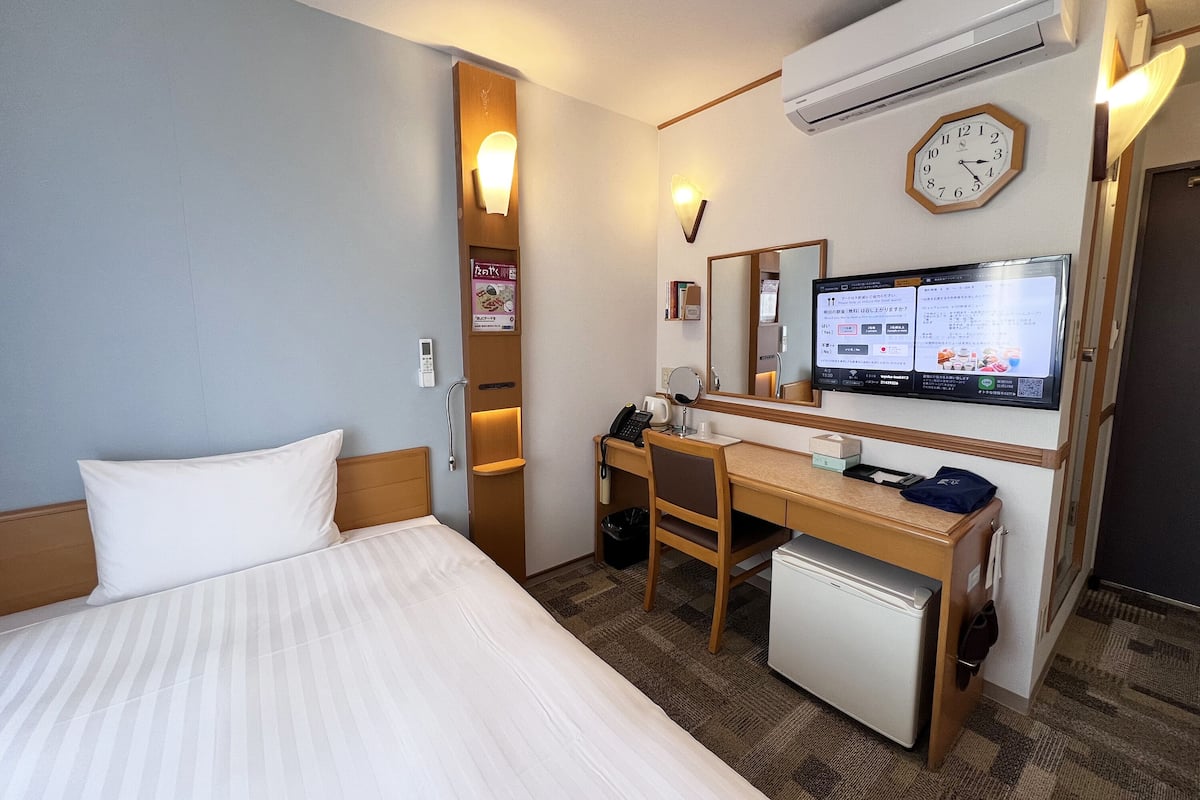 Une chambre d’hôtel équipée d’un lit, d’un bureau, d’un petit réfrigérateur, d’un miroir, d’une horloge et d’un écran numérique affichant des informations.