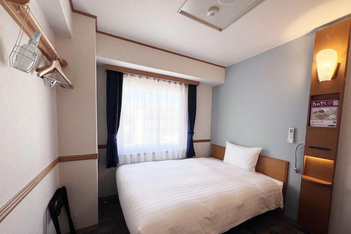 Une chambre d’hôtel avec un grand lit, une fenêtre avec des rideaux, une table de chevet avec une lampe et un téléviseur fixé au mur.