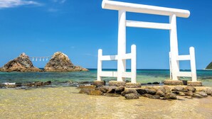 A white torii gate at a beach with clear blue water and rocky outcrops in the background.