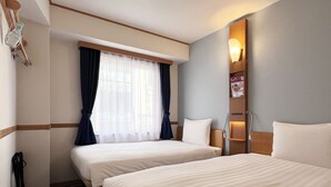 A hotel room with two beds, a window with curtains, a bedside table, and a wall-mounted lamp.