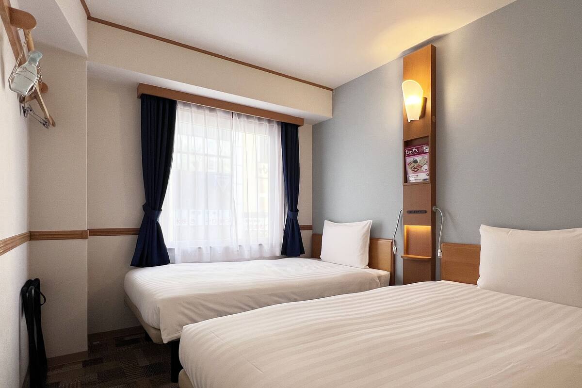 Une chambre d’hôtel avec deux lits, une fenêtre avec des rideaux, une table de chevet et une lampe fixée au mur.