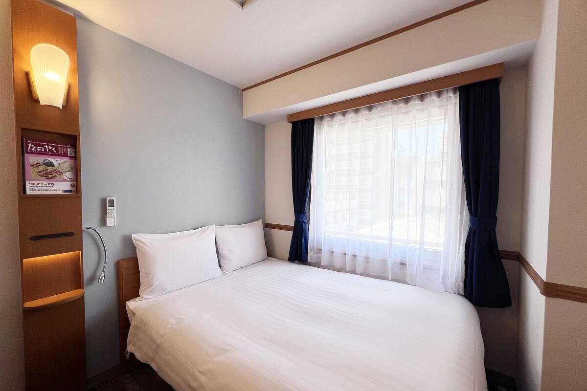 Une chambre d’hôtel avec un grand lit, une table de chevet, une lampe fixée au mur et une fenêtre avec des rideaux.