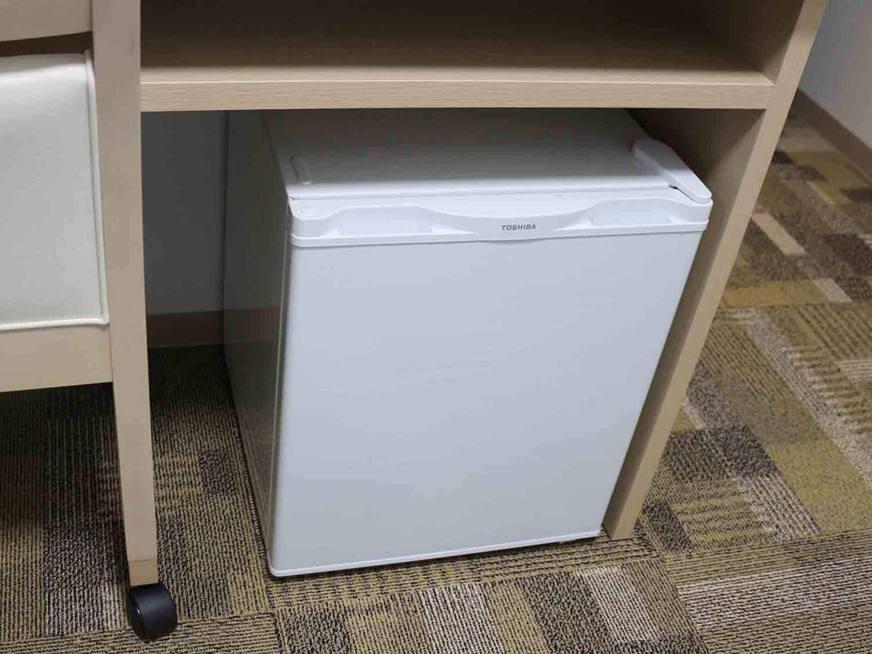 A white Toshiba refrigerator under a wooden desk with a carpeted floor.