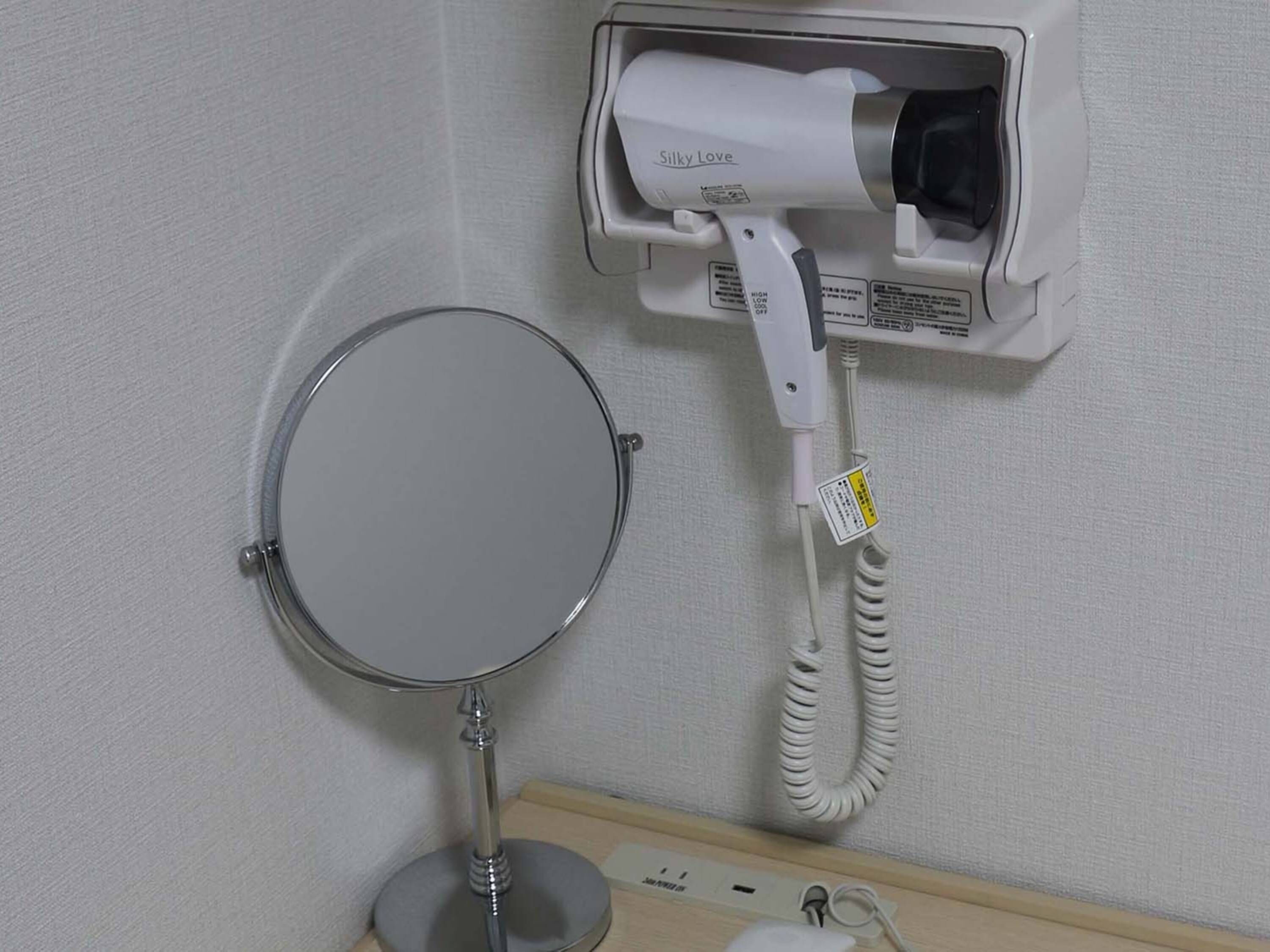 Ein an der Wand befestigter Haartrockner, ein runder Spiegel auf einem Ständer und eine Steckdose mit Stecker.