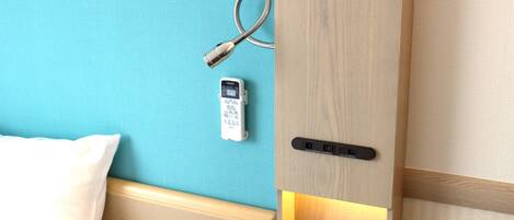 A hotel room with a wooden headboard, a bedside table, a clock, a telephone, and a power outlet.