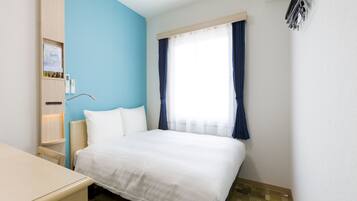 A hotel room with a bed, a desk, a nightstand, a wall-mounted shelf with toiletries, and a window with curtains.