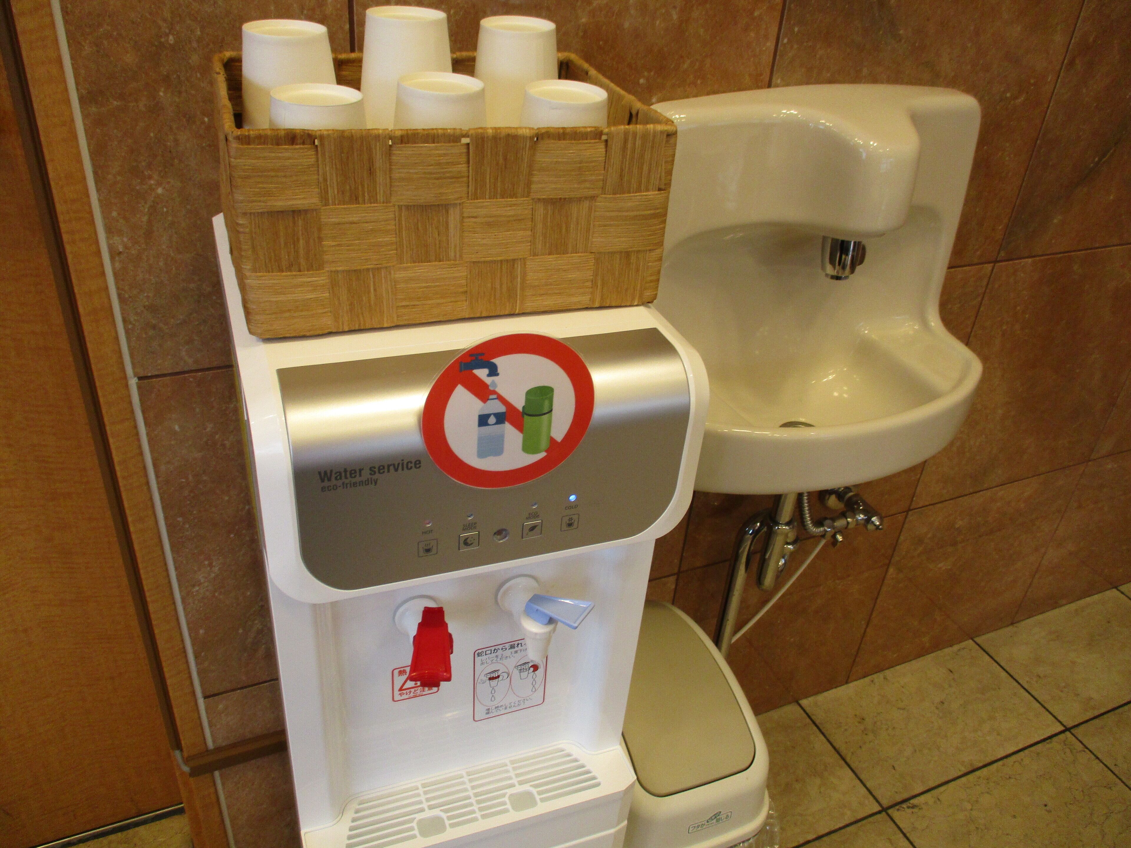 Un distributore d'acqua con un rubinetto a funzionamento senza contatto, un pulsante rosso e un pulsante blu, e un contenitore di legno con asciugamani di carta.