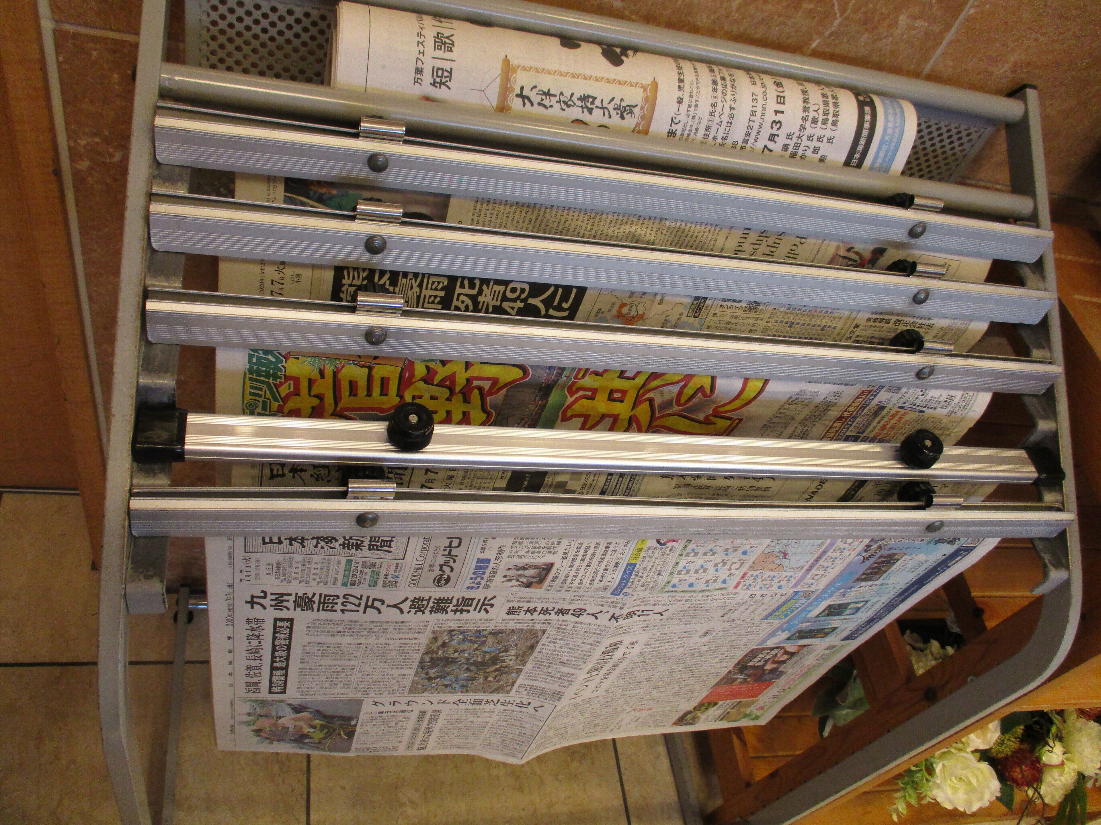 A metal rack with newspapers and magazines, some with Japanese text, and a wooden table with flowers.