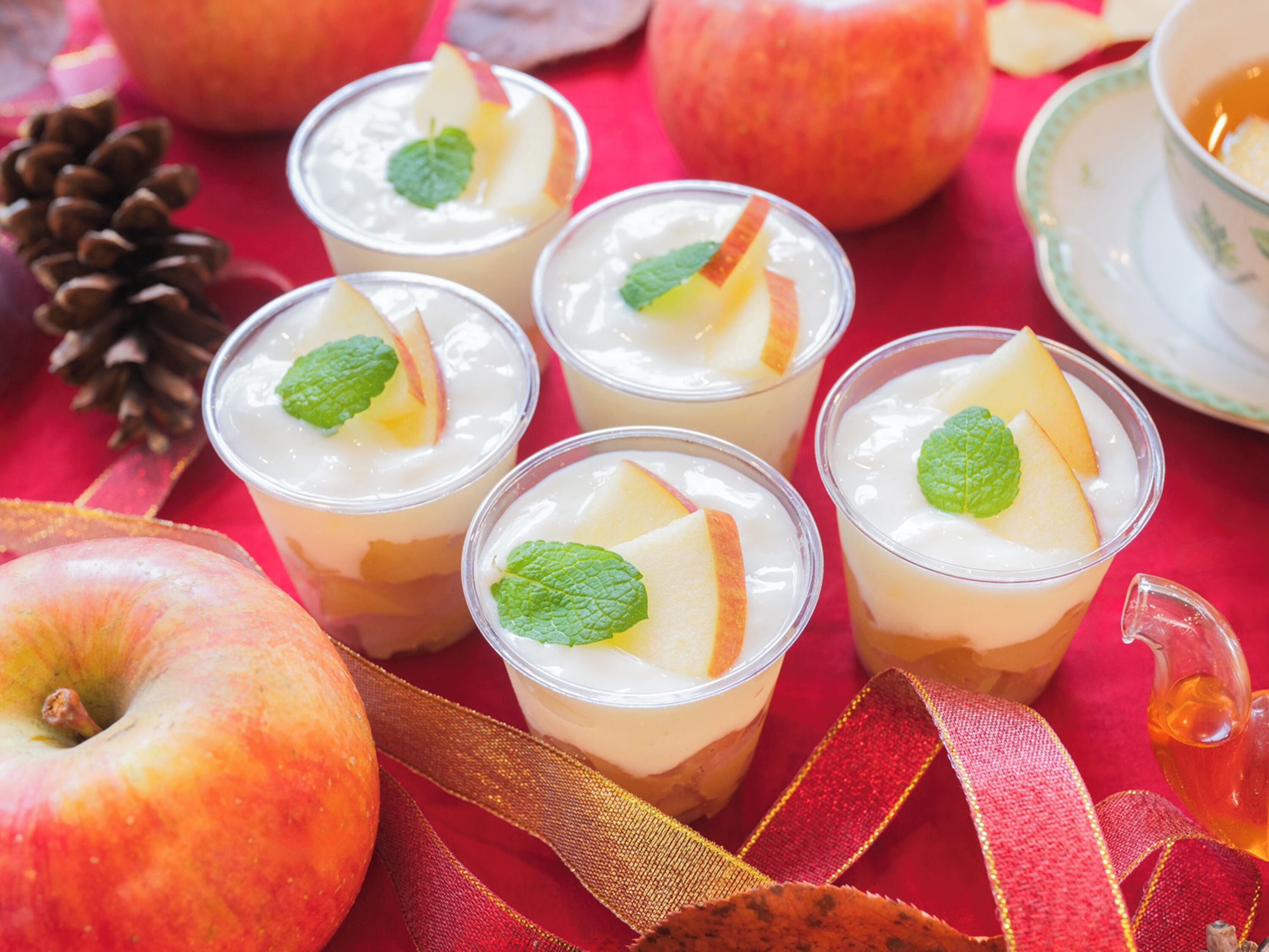 Cuatro vasos de postre con rodajas de manzana y hojas de menta, rodeados de manzanas y un lazo decorativo sobre un mantel rojo.